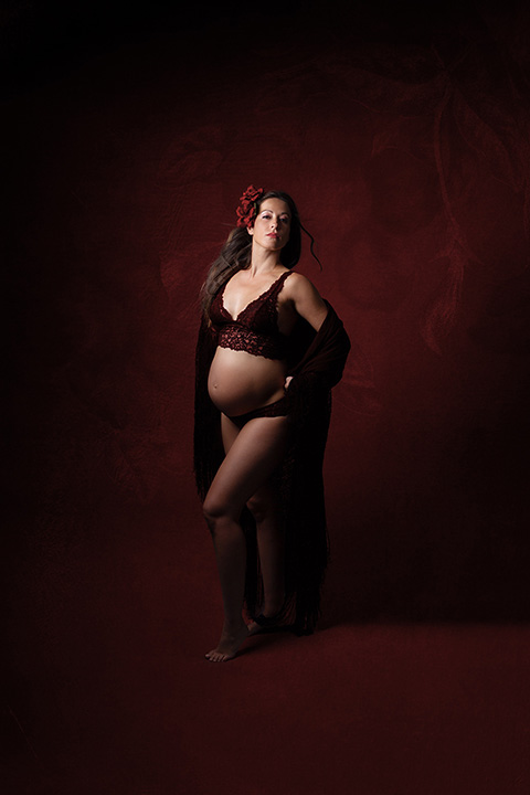 Fotografía de embarazo estilo flamenco, la chica está de pie, vestido con ropa interior roja de encaje, flor en la cabeza y sujeta con sus codos un mantón negro.