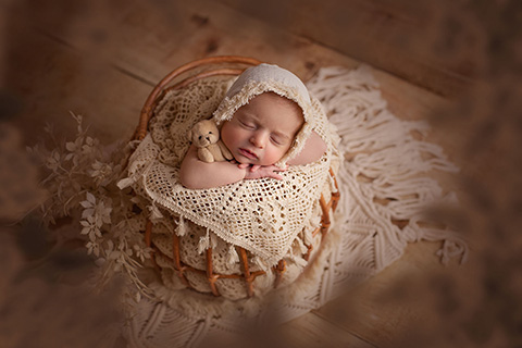 Bebé recién nacido metido en una cesta de ratán, lleva un gorro blanco y está sobre una manta de encaje, además sujeta un oso pequeño de peluche en su brazo.