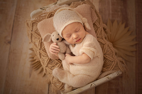 Bebé con 10 días de vida, dormido sobre una cama blanca, está boca arriba con las piernas flexionadas y sujetando un conejo de peluche con sus manos, además lleva un body y un gorro dormilón en tono beige.
