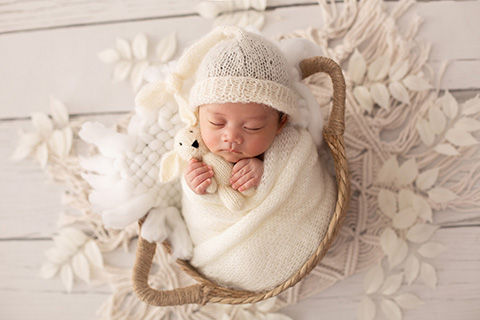 Sesión newborn con un bebé sobre una cesta rodeado de hojas blancas, lleva un gorro dormilón y sujeta un conejo entre sus manos.