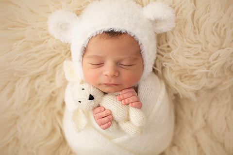 Bebé newborn dormido sobre una manta de pelo blanca, envuelto con una tela del mismo color, además lleva un gorro con forma de oso.