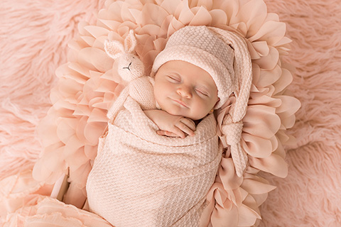 Niña dormida y envuelta en una tela rosa, solo se le ven las manos, lleva un gorro dormilón que cuelga hacia abajo, entre sus manos está agarrando un conejo de peluche rosa, la bebé está tumbada sobre una manta de tul y de pelo, imagen realizada por fotógrafo newborn Madrid.