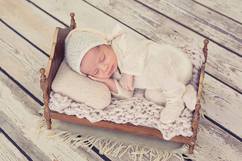 Bebé dormido de 14 días tumbado sobre una camita muy pequeña de madera, tiene una manta y un cojín en color beige, su mano está apoyada sobre su mejilla, está en un estudio en sus fotografías de newborn Madrid.