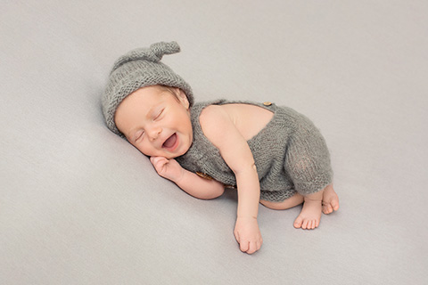 Imagen de un bebé dormido, está tumbado sobre una manta gris, sonríe, lleva un gorro de lana gris y un peto a juego en su reportaje de recién nacido.
