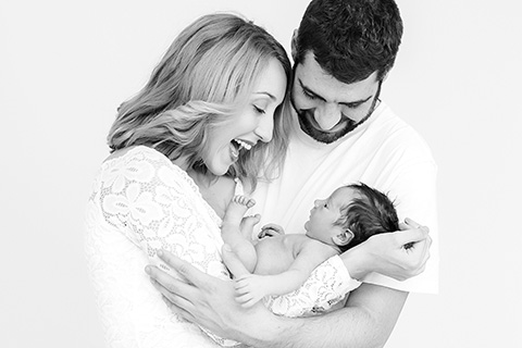 Padres abrazando a su bebé, le sonríen, es una imagen en blanco y negro, llevan camisetas blancas y la niña está sin ropa, imagen que realizaron fotógrafos profesionales de newborn.