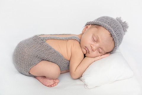Bebé dormido sobre un cojín una manta blanca, lleva un gorro con un pompón y un peto de lana, imagen realizada en unas sesiones de newborn en Madrid.