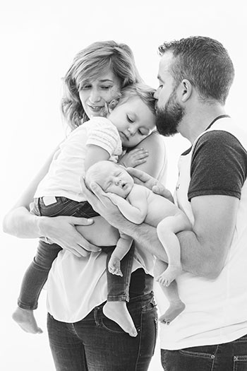 Padres cogiendo a sus dos hijas en brazos, el padre le está dando un beso en la frente a la mayor, están abrazados y visten camisetas blancas en un reportaje familiar en Madrid.