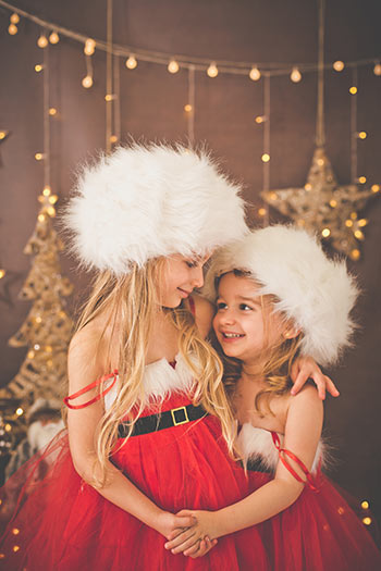 Dos hermanas se abrazan y se miran en una sesión de fotos infantil, llevan ropa navideña, un gorro ruso blanco de mucho pelo y un vestido rojo de mamá Noel, el fondo tiene luces y estrellas.